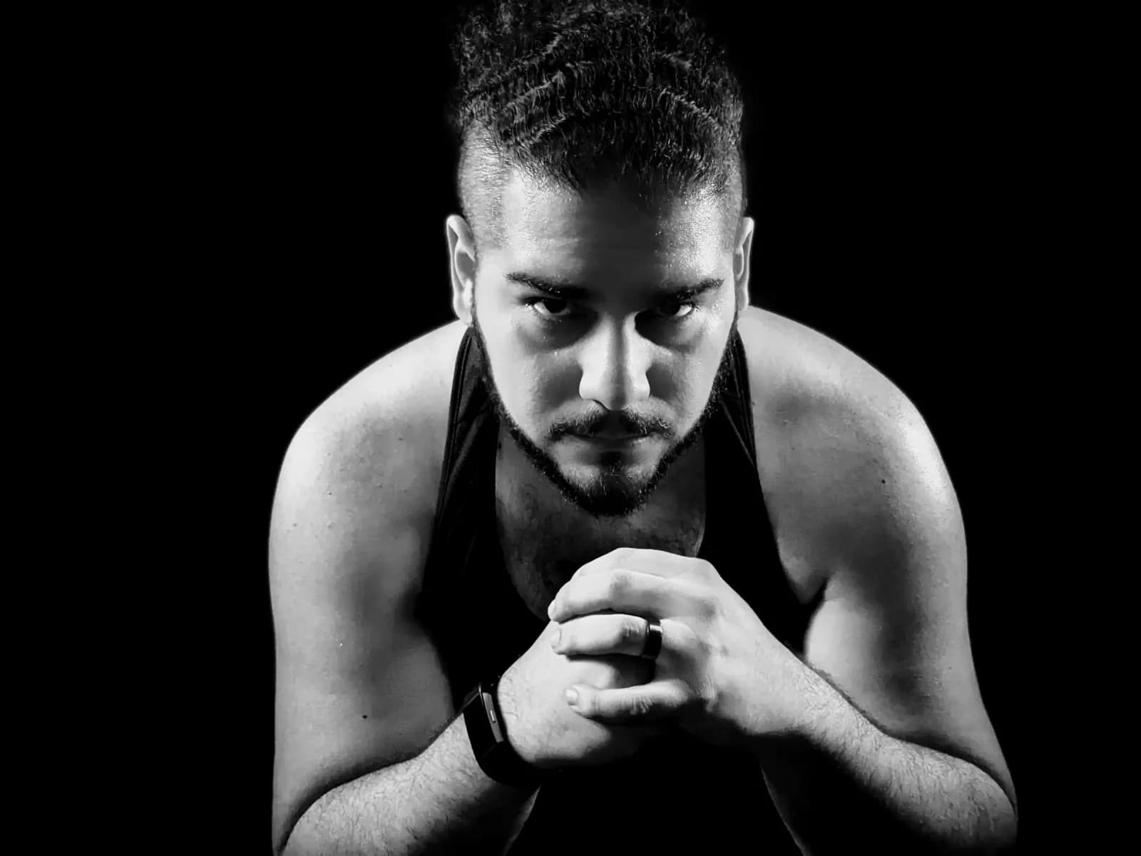 Black-and-white portrait of a lifter in a tank top, leaning forward with hands clasped, looking straight at the camera